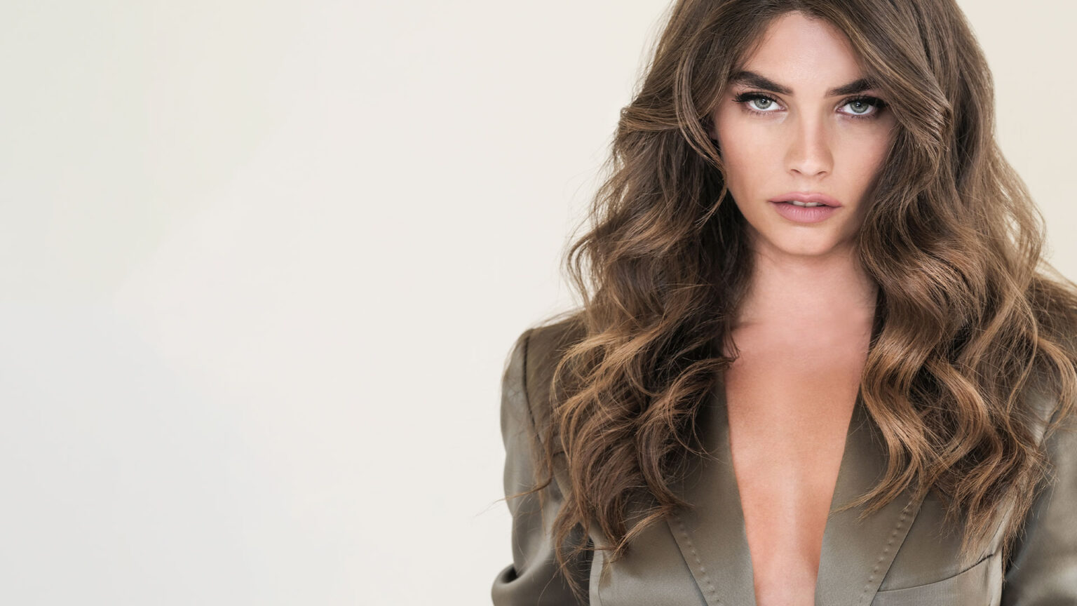 Portrait of a woman with long wavy brown hair wearing an olive blazer, looking directly at the camera.
