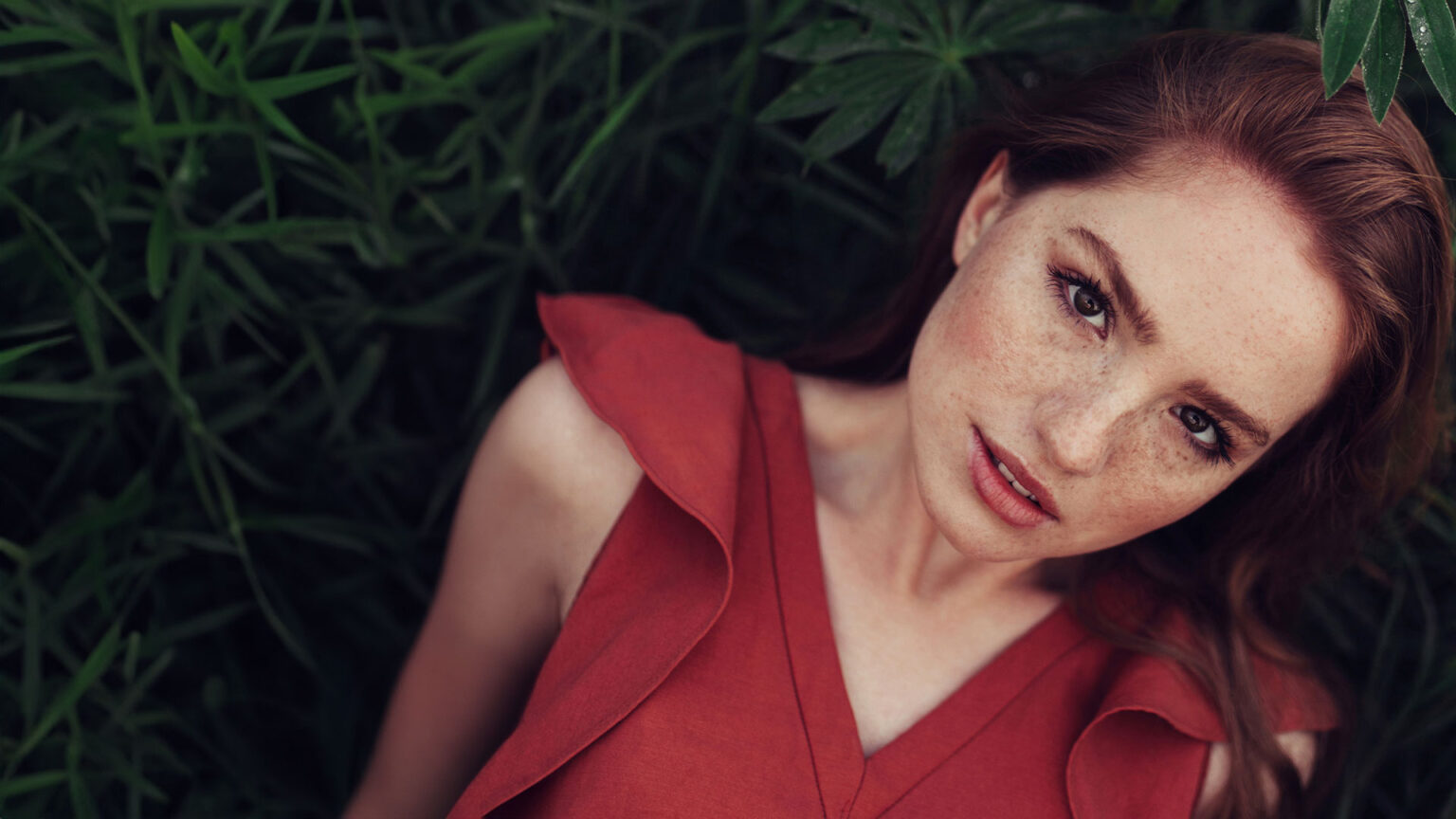 Portrait of a woman with freckles and long auburn hair, lying among green foliage while wearing a red dress.