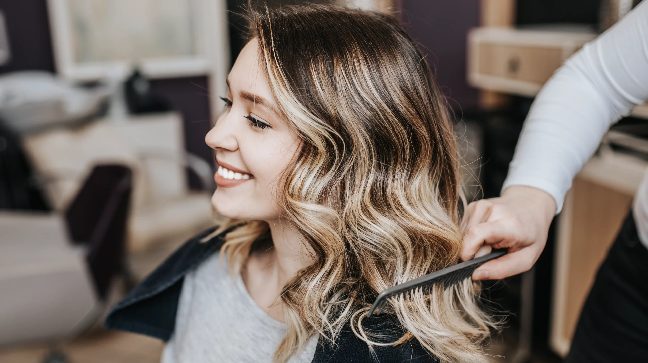 Smiling model receiving essential-level highlights, with soft balayage tones being refined by a stylist using a comb in a modern salon setting