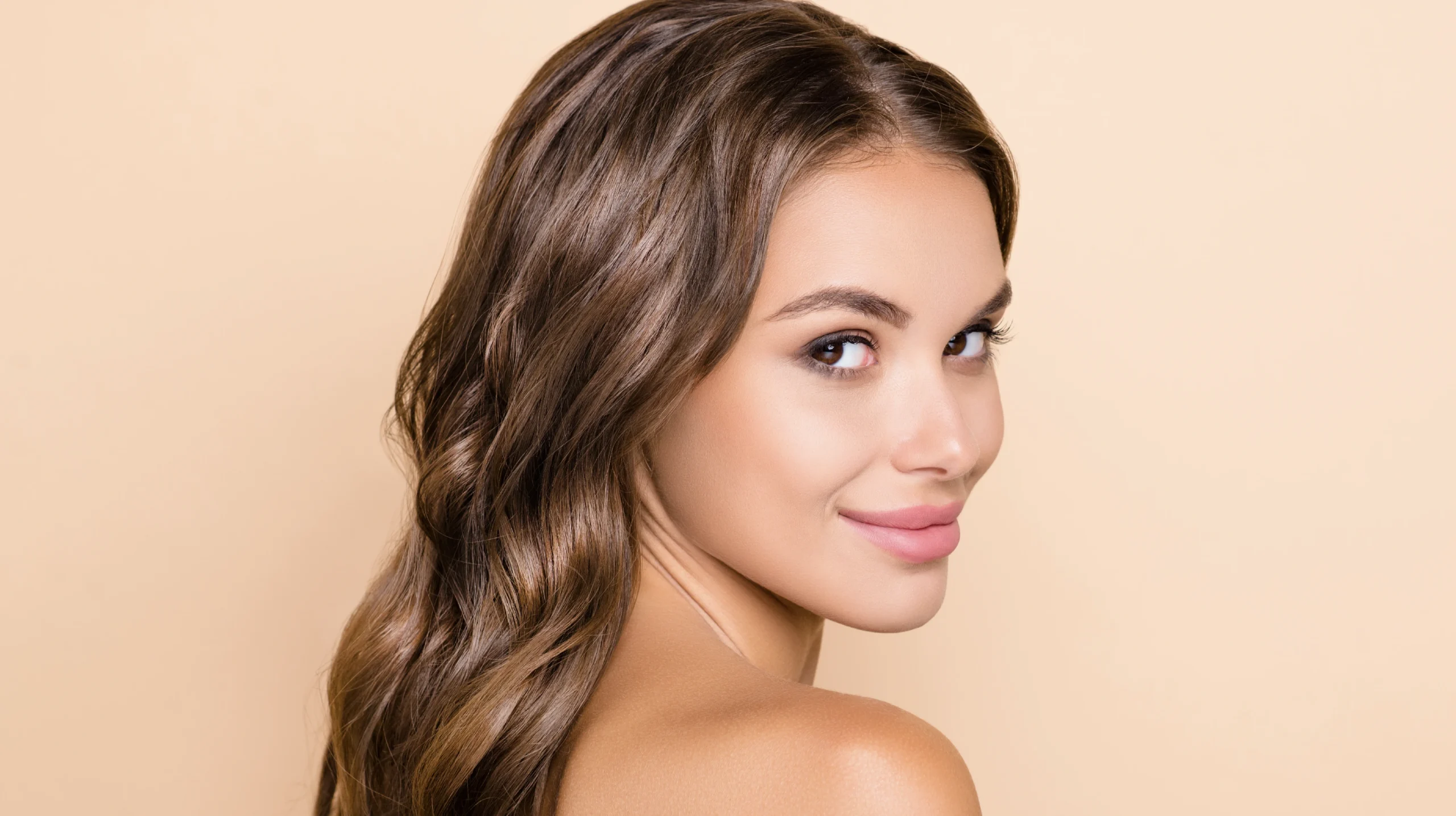 Three-quarter profile of a model with softly waved brown hair and natural color balance, showing smooth texture and even shine against a warm neutral background