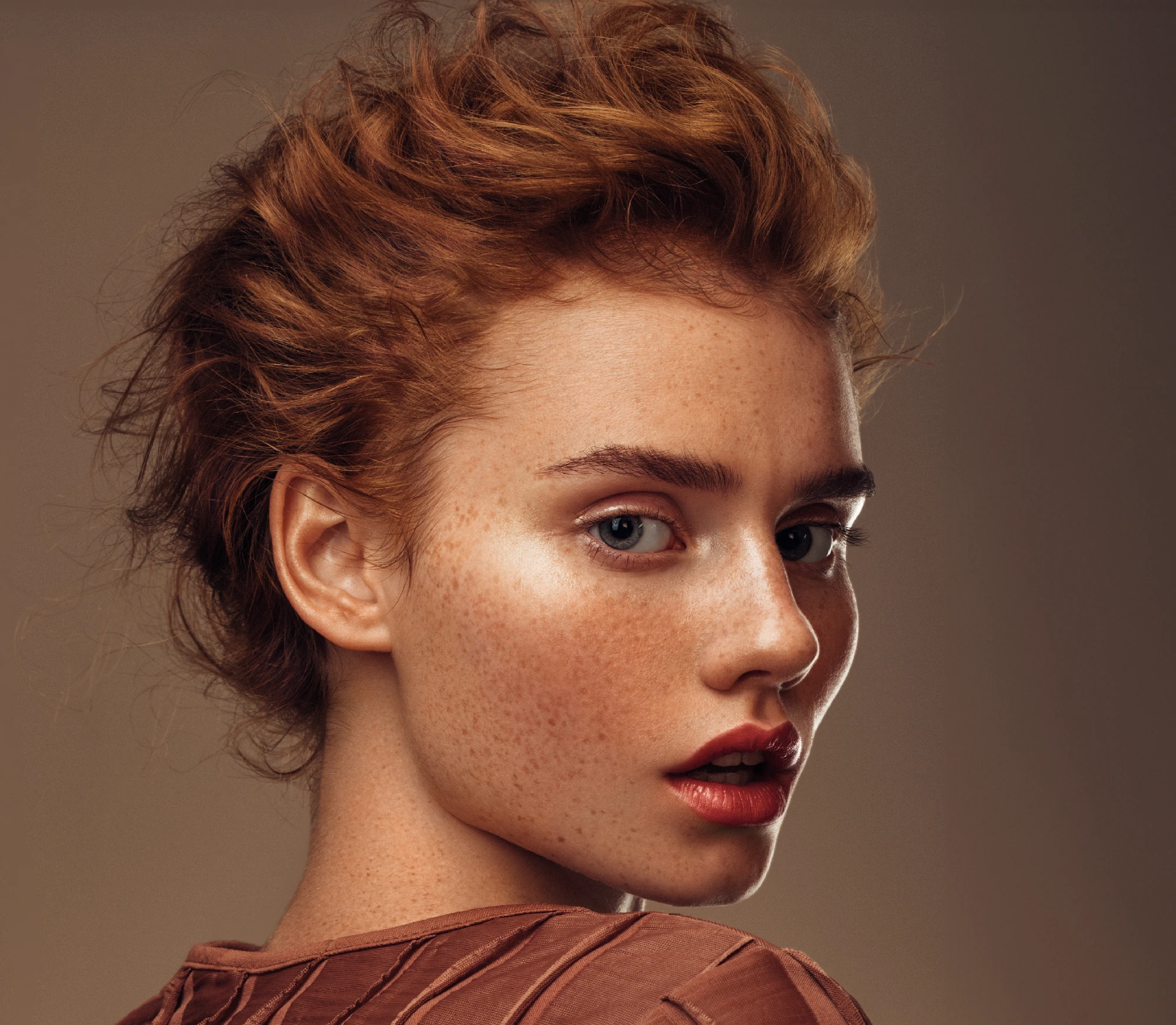 Portrait of a woman with copper-red hair, fair freckled skin, and natural makeup, photographed against a warm neutral background.