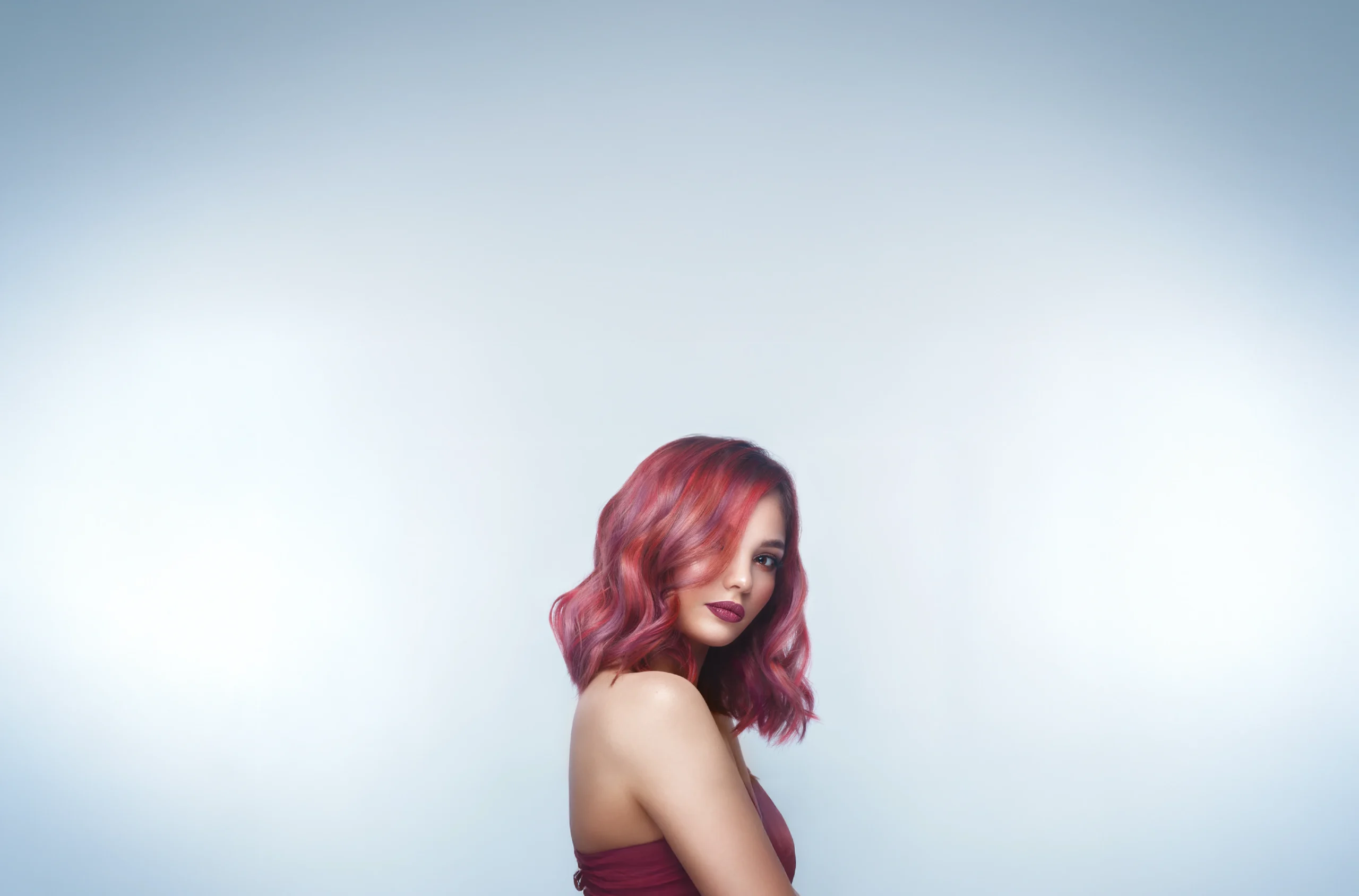 Female model posing in profile with shoulder-length wavy pink hair, wearing a sleeveless top against a light gradient studio background.