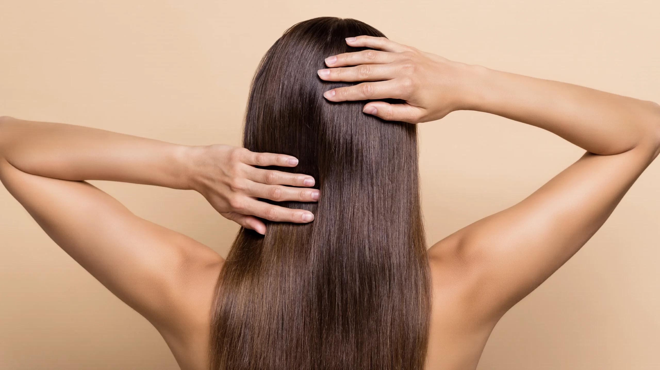 Back view of a woman with long straight shiny hair touching her scalp with both hands on a neutral background