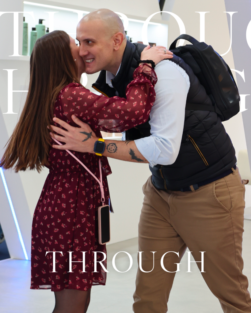 Man and woman greeting each other with a cheek kiss while embracing at an indoor event, with product shelves in the background.