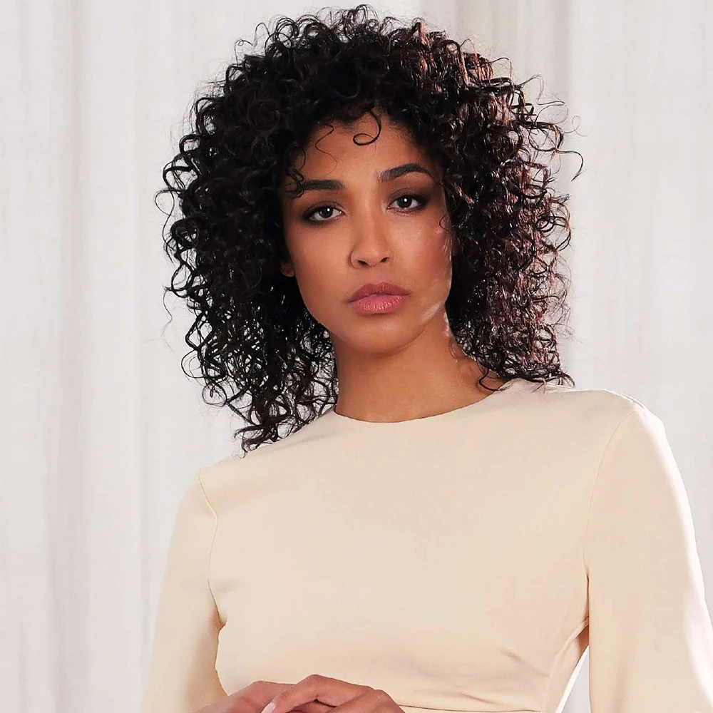 Model with dark shoulder-length curls and an ivory long-sleeved top poses with hands together against pale curtains.