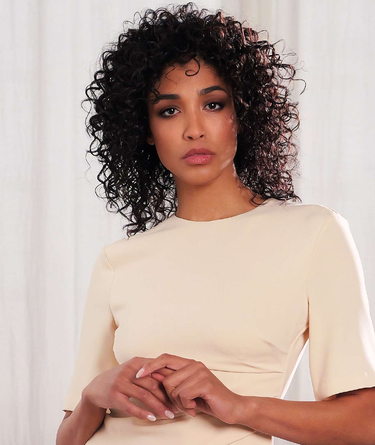 Cocoa Muse portrait with dark shoulder-length curls and a cream top against pale curtains.
